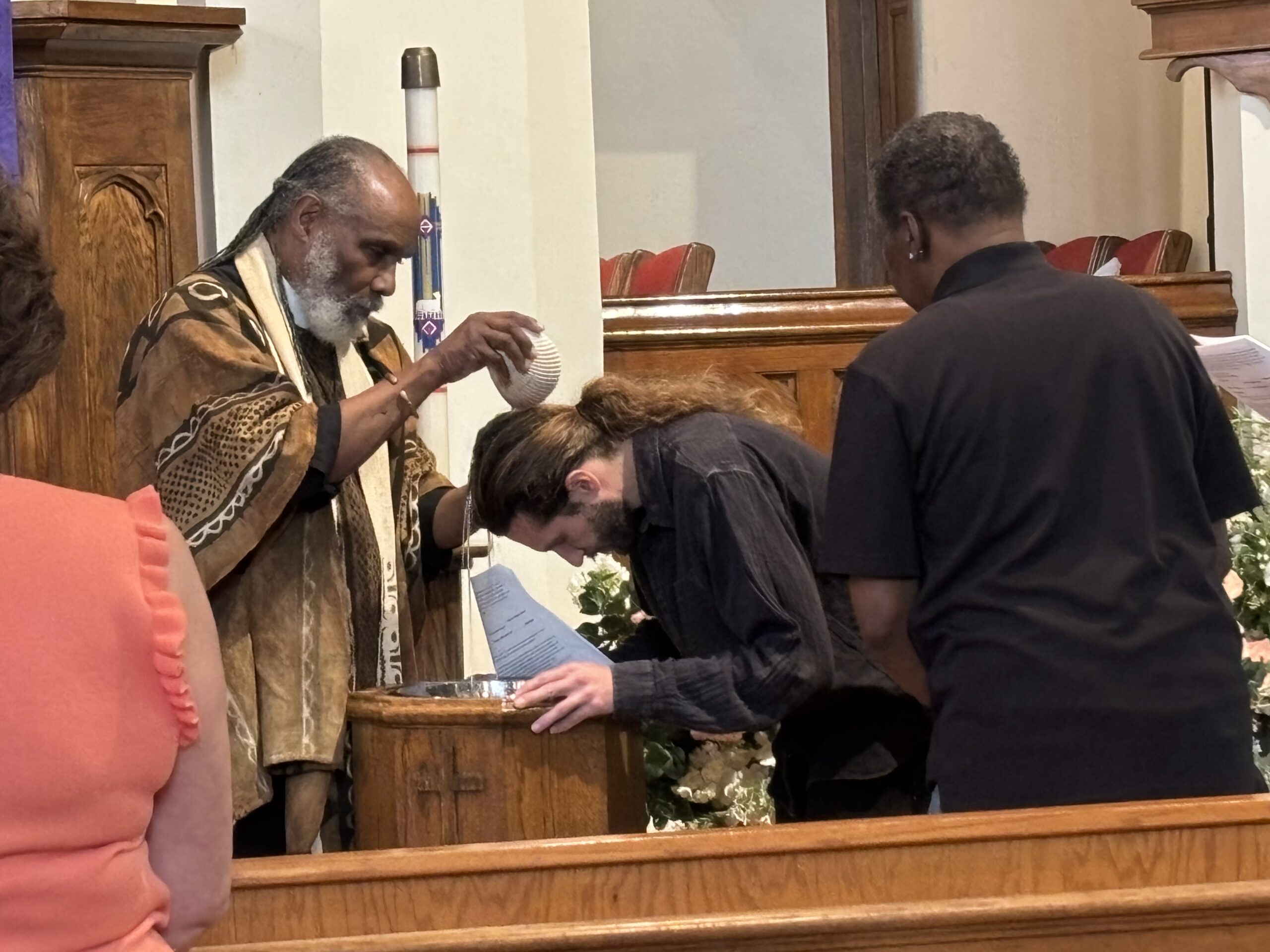 Pastor Starr pours water from a ceramic bowl over the back of the head of an adult male congregant with a ponytail and a beard. The congregant is leaning his head over the baptismal font which he is holding with both hands.